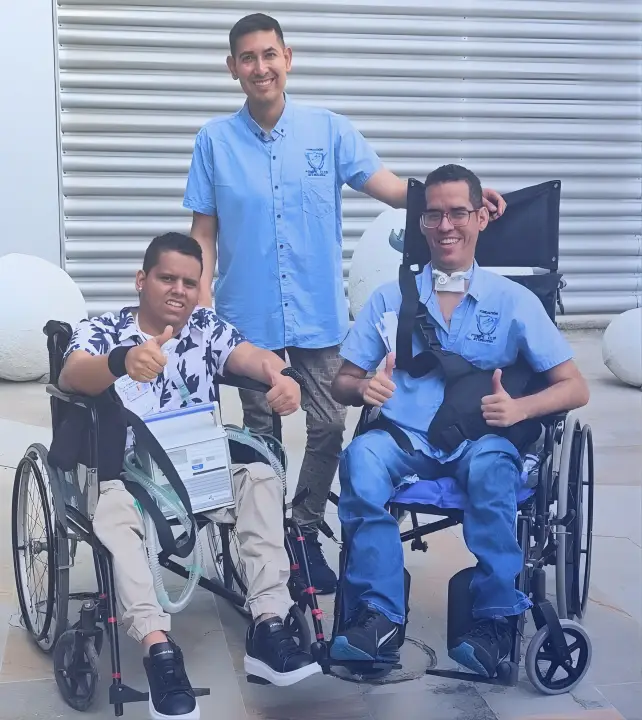 Brayhan González y Fernando Rodríguez, líderes de la Fundación Pompe Club Internacional, posan sonrientes junto al miembro de la comunidad Miller. Fernando está de pie en el centro, apoyando sus manos sobre los hombros de Brayhan y Miller, quienes están sentados en sus sillas de ruedas haciendo un gesto de aprobación con los pulgares arriba.
