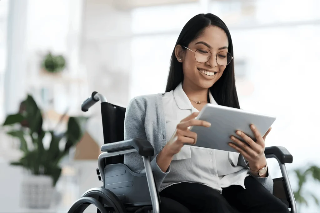 Mujer joven con gafas y cabello oscuro sonriendo mientras utiliza una tableta electrónica. Está sentada en una silla de ruedas en una habitación luminosa y moderna con plantas de fondo, transmitiendo una imagen de autonomía y conectividad digital.